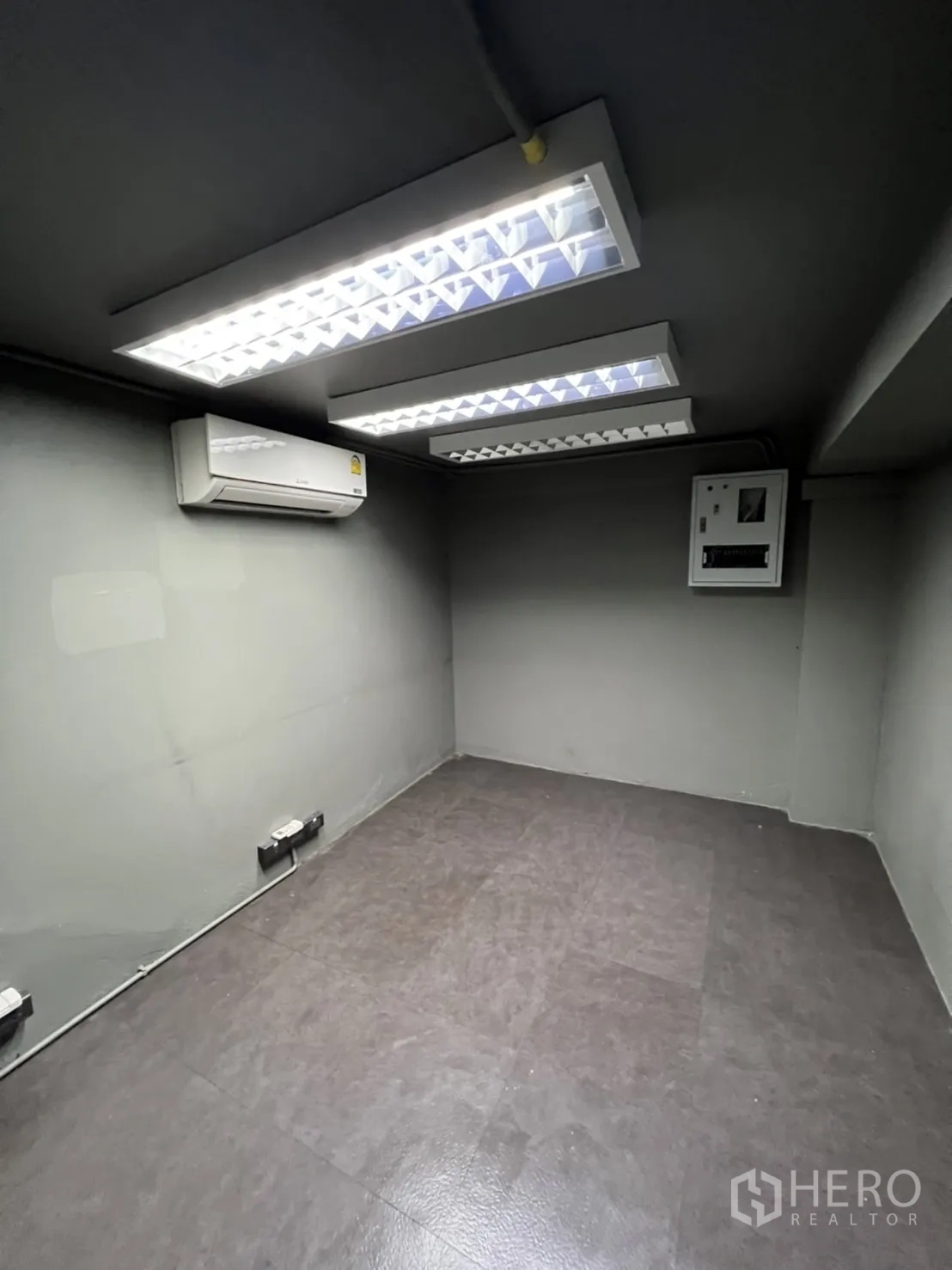 Shophouse for rent in Phra Khanong, Bangkok - Compact office room with fluorescent ceiling lights, split air conditioner and an electrical panel on grey walls.
