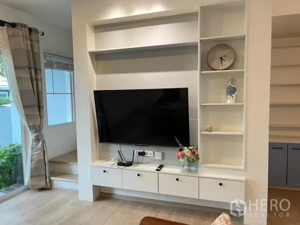 Townhouse for rent in Prawet, Bangkok - Living room with built‑in media wall, shelving, flat‑screen TV, and light wood flooring.