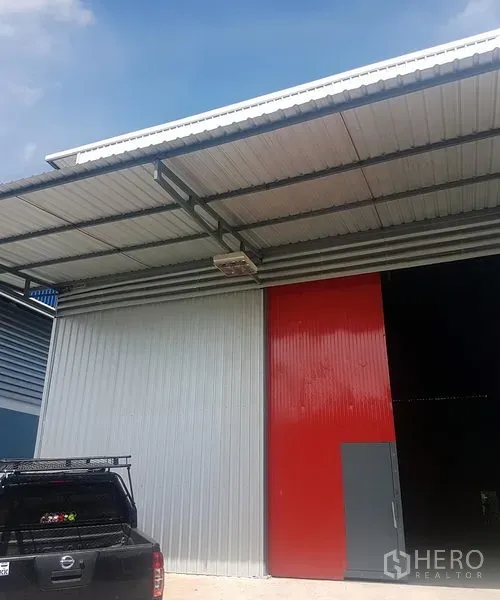 Warehouse for rent in Prawet, Bangkok - Exterior facade of the warehouse with red and gray metal sliding door, canopy and parking area.