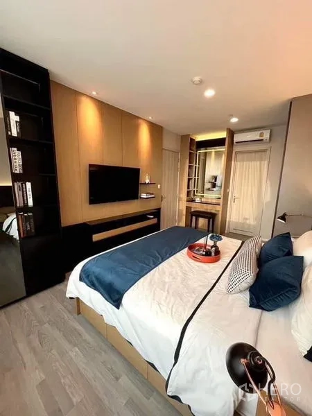 Condominium for sale in Bang Sue, Bangkok - Bedroom with wall-mounted TV, built-in wardrobe and vanity, warm wood flooring and downlights.