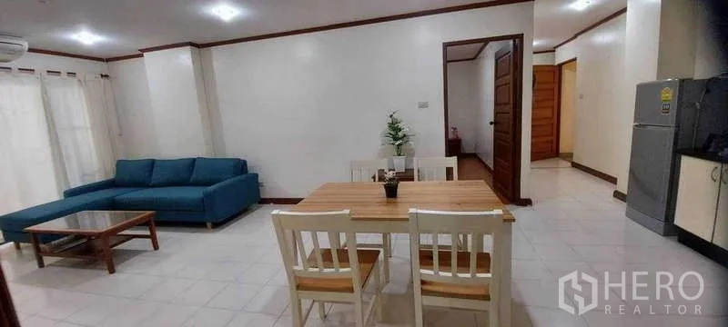 Condominium for rent in Khlong Toei, Bangkok - Dining and living space with a four‑seat wooden table, blue sofa, tile floors and sheer balcony curtains.
