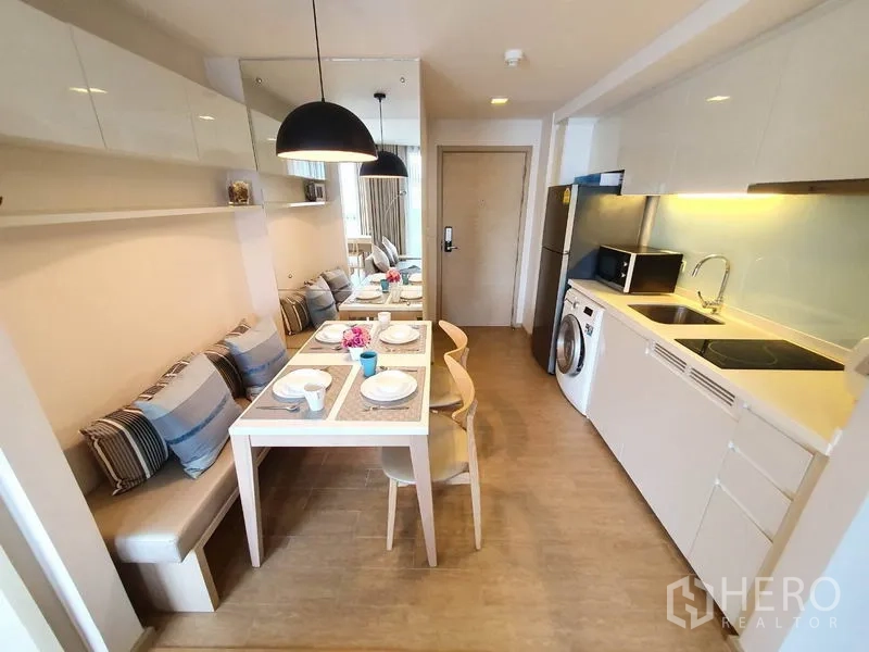 Condominium for rent in Watthana, Bangkok - Compact modern kitchen with white cabinets, sink, cooktop, microwave and in-unit washing machine next to the dining area.