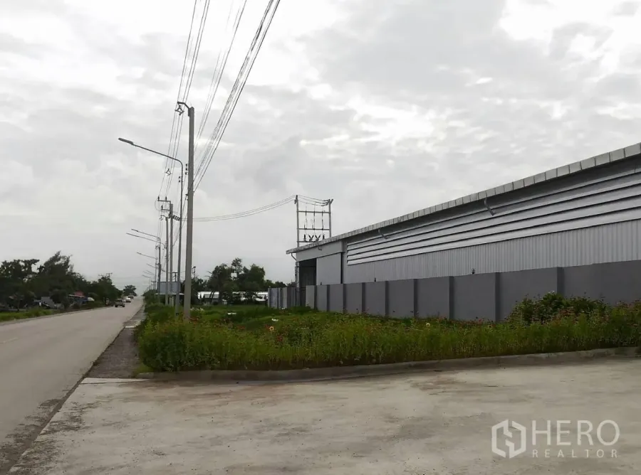 Warehouse for rent in Lam Luk Ka, Pathum Thani - Street-side view of a modern gray warehouse in Lam Luk Ka with a perimeter wall, power lines and a wide concrete entry.