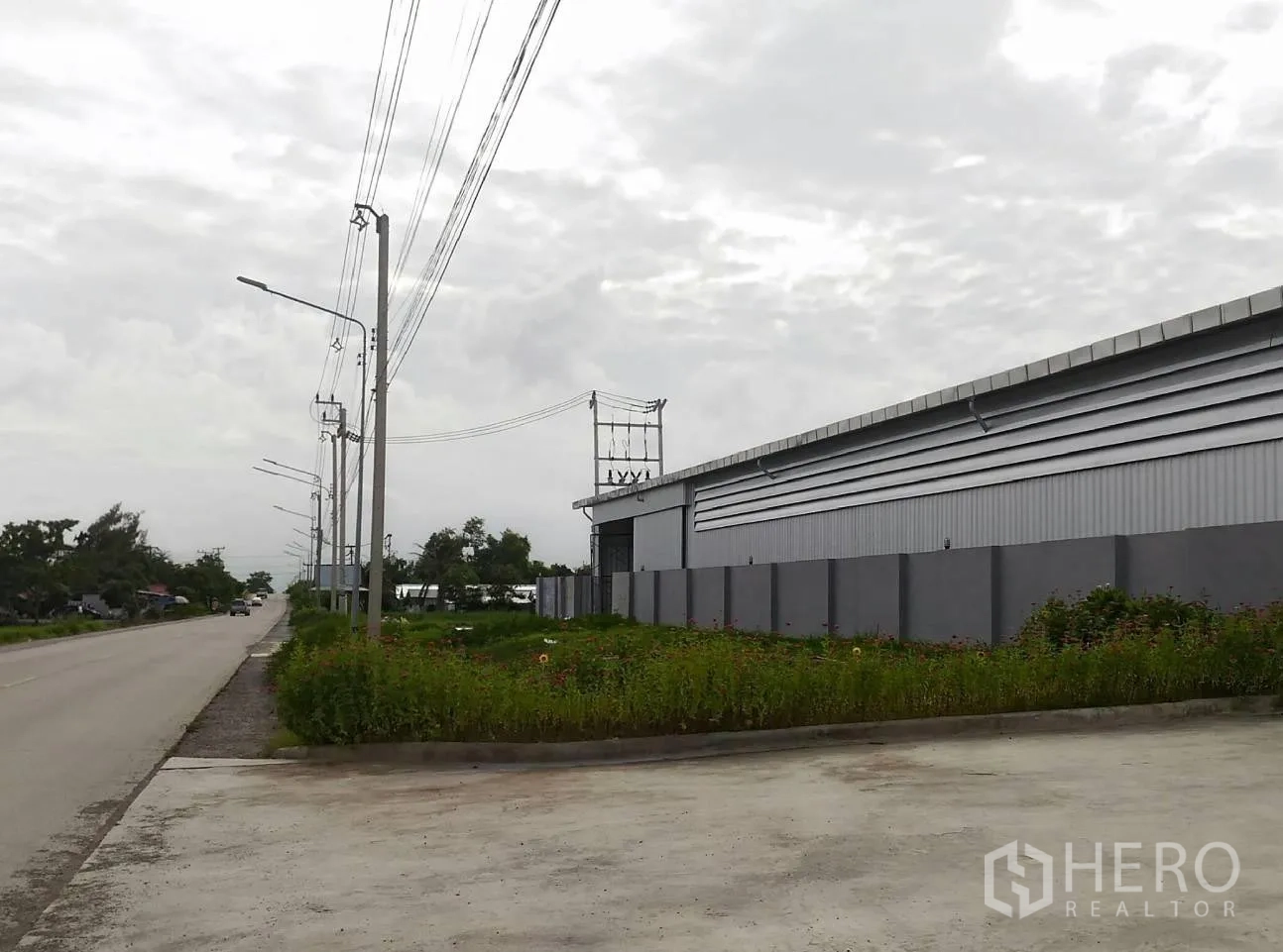 Warehouse for rent in Lam Luk Ka, Pathum Thani - Street-side view of a modern gray warehouse in Lam Luk Ka with a perimeter wall, power lines and a wide concrete entry.