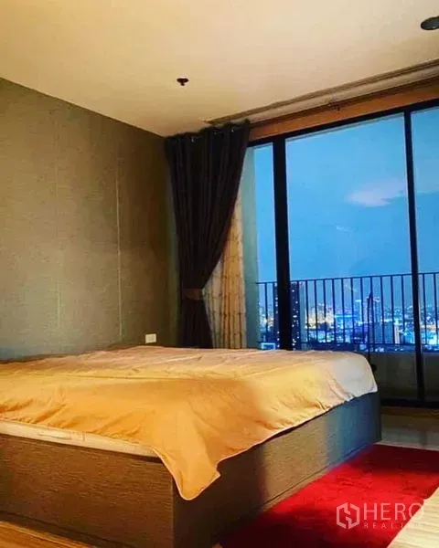 Condominium for rent in Watthana, Bangkok - Cozy bedroom with large window, blackout curtains and night city view from a high floor balcony.
