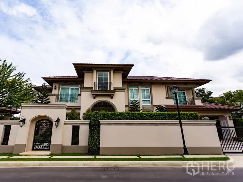 Detached House for rent in Mueang Samut Prakan, Samut Prakan - Two‑storey detached house with high perimeter wall, iron gate and manicured hedges on a quiet street.