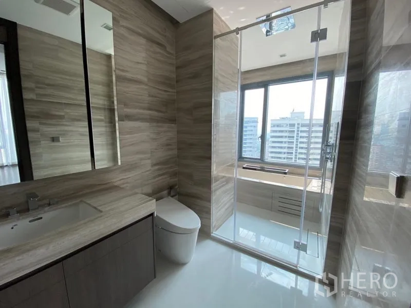 Condominium for rent in Khlong Toei, Bangkok - Contemporary bathroom with stone-textured walls, glass shower and a wide window facing city buildings.
