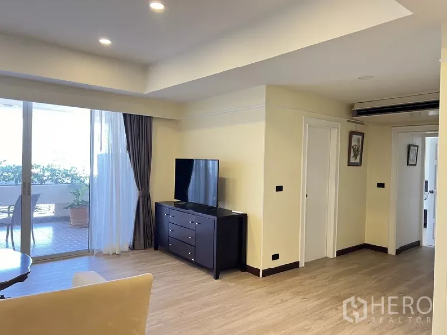Condominium for rent in Khlong Toei, Bangkok - Open-plan lounge with recessed lights, wood floors and access to a large covered balcony beside a TV stand.