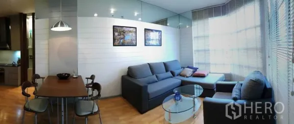 Condominium for rent in Khlong Toei, Bangkok - Bright living room with L-shaped sofa, glass coffee table and large window blinds.