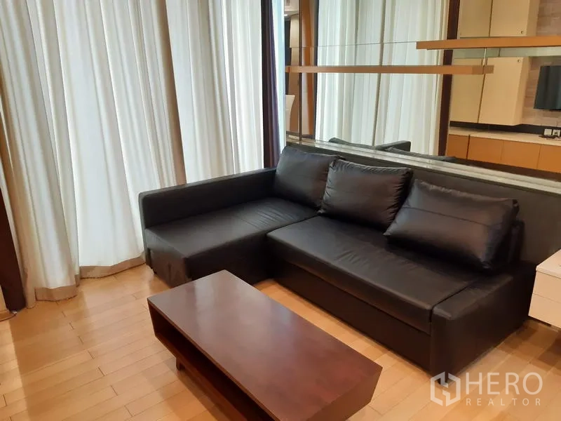Condominium for rent in Khlong Toei, Bangkok - Cozy living room with a black L-shaped sofa, wooden coffee table, mirrored wall, and floor-to-ceiling curtains.