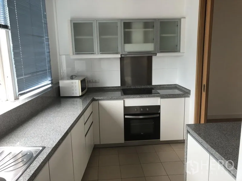 Condominium for rent in Khlong Toei, Bangkok - Enclosed modern kitchen with L-shaped granite counters, glass-front cabinets and built-in oven.