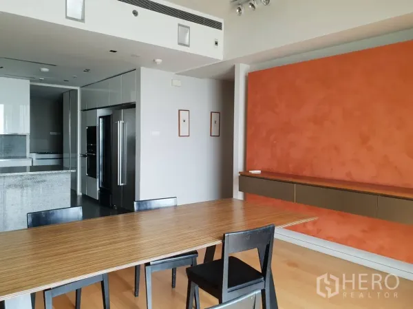 Condominium for rent in Sathon, Bangkok - Dining space facing the sleek kitchen, with a wood table, black chairs and a textured orange accent wall in a modern condo.