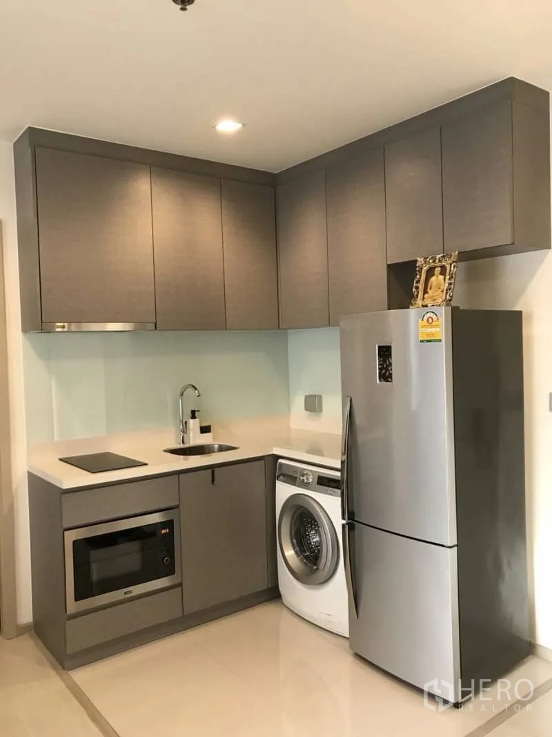 Condominium for rent or sale in Khlong Toei, Bangkok - Compact built-in kitchen with cabinets, cooktop, fridge and washing machine.
