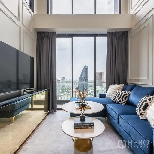 Condominium for rent in Khlong Toei, Bangkok - Lounge area with blue sofa, coffee tables and TV facing panoramic city views through full-height windows and a high ceiling.