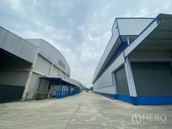 Warehouse for rent in Pathum Thani - Wide concrete driveway between two modern steel warehouses with loading bays and roller shutter doors.