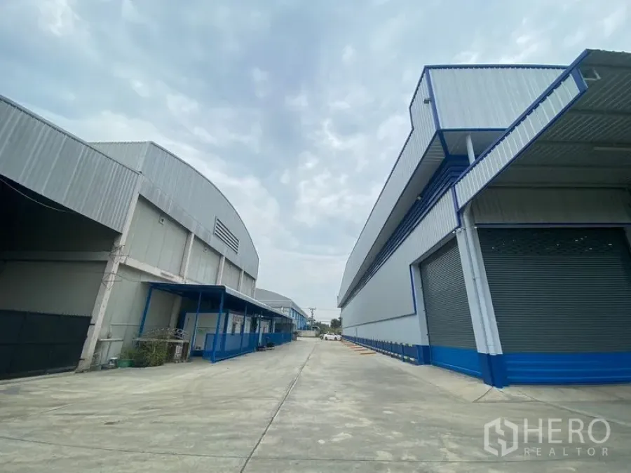 Warehouse for rent in Pathum Thani - Wide concrete driveway between two modern steel warehouses with loading bays and roller shutter doors.