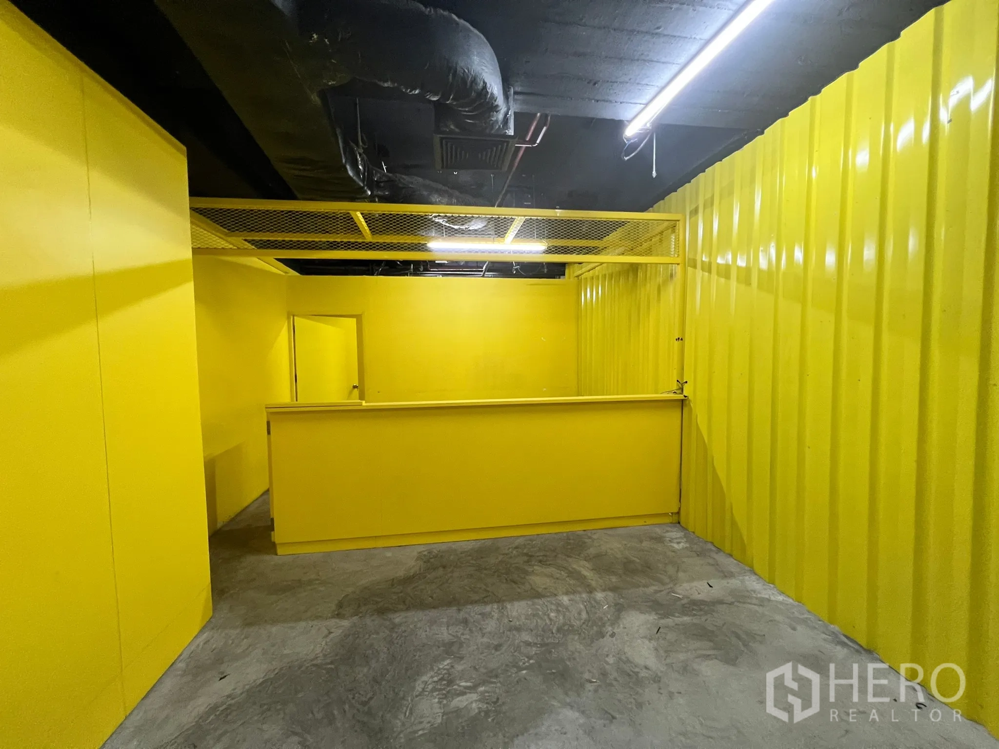 零售空间 出租 - 接待吧台区，黄色波纹墙、金属网格吊顶与日光灯照明。