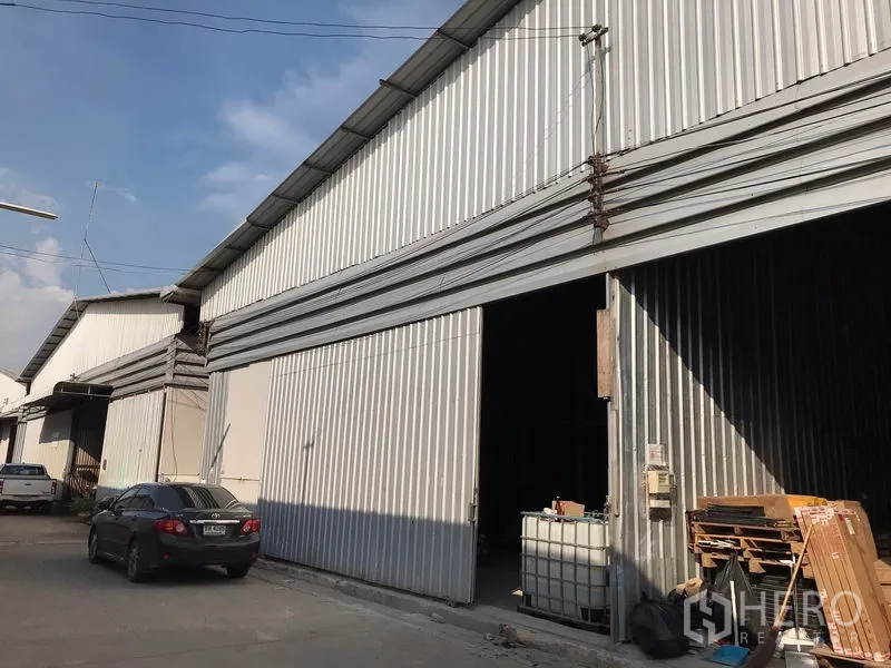 Warehouse for rent in Mueang Samut Prakan, Samut Prakan - Metal-clad warehouse exterior with wide sliding doors, driveway parking and overhead power lines along an industrial lane in Mueang Samut Prakan.
