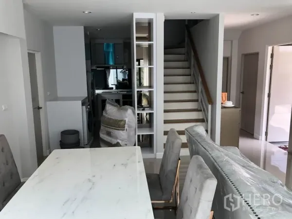 Detached House for rent in Prawet, Bangkok - Open-plan dining area with a marble table facing a staircase and built-in shelving.