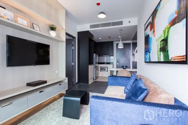 Condominium for rent in Watthana, Bangkok - Open-plan living room with blue sofa, TV wall and compact kitchen in a Watthana condo.