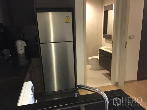 Condominium for rent or sale in Watthana, Bangkok - View of a stainless-steel refrigerator and doorway to a contemporary bathroom from the kitchen with black countertop.