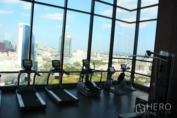Condominium for rent in Huai Khwang, Bangkok - High-rise fitness center with treadmills and cardio machines facing the Bangkok skyline through floor-to-ceiling windows.