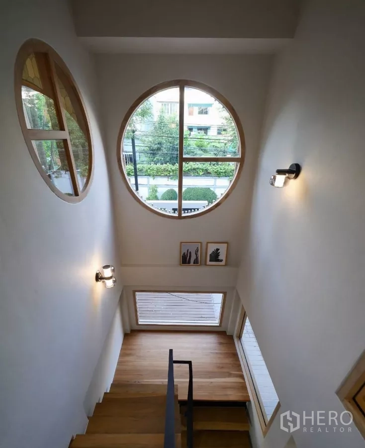 Townhouse for rent in Watthana, Bangkok - Stair landing with a large round window and wooden steps overlooking greenery at the front.