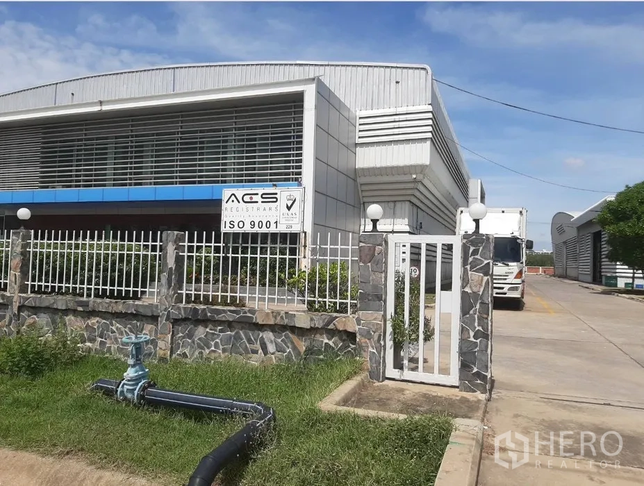 Factory for rent or sale in Uthai, Phra Nakhon Si Ayutthaya - Modern factory exterior with louvered facade, blue canopy, gated fence and a wide driveway with a truck.