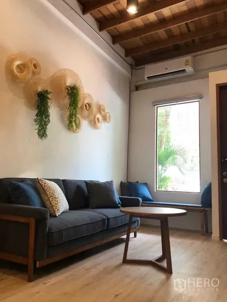 Detached House for rent in Phra Nakhon, Bangkok - Cozy living room featuring a gray sofa, wooden ceiling beams, wall art, and a window with garden view.