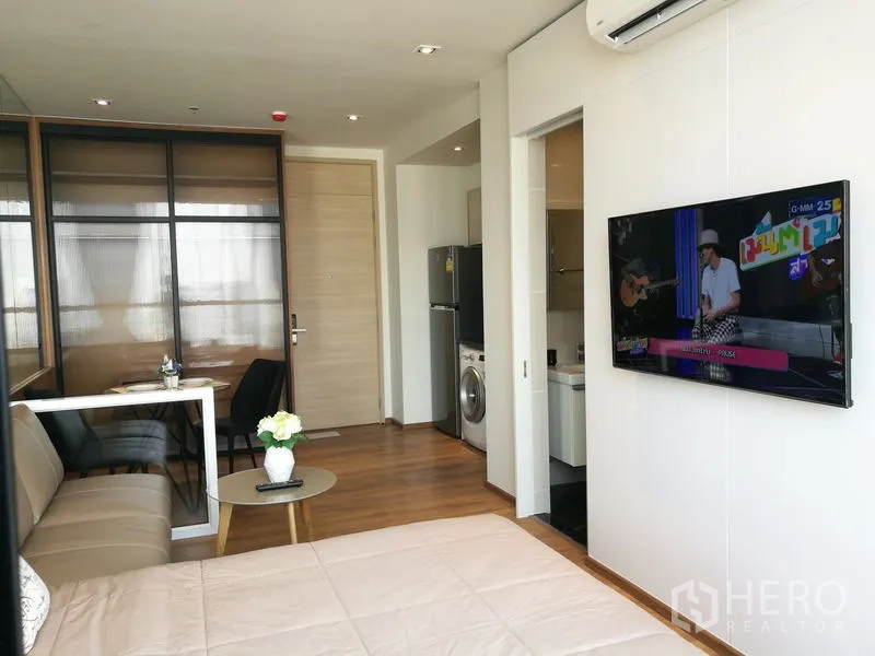 Condominium for sale in Khlong Toei, Bangkok - Wall-mounted TV opposite the bed with entryway and modern kitchenette in view.