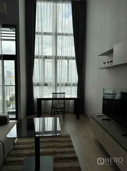 คอนโด ให้เช่าในHuai Khwang, Bangkok - ห้องนั่งเล่นสว่างด้วยผ้าม่านสูง โต๊ะทำงาน ตู้วางทีวี และวิวเมืองกว้าง