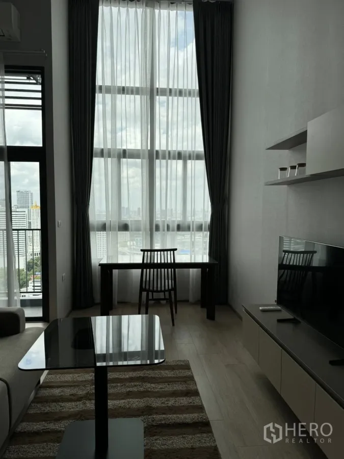 คอนโด ให้เช่าในHuai Khwang, Bangkok - ห้องนั่งเล่นสว่างด้วยผ้าม่านสูง โต๊ะทำงาน ตู้วางทีวี และวิวเมืองกว้าง