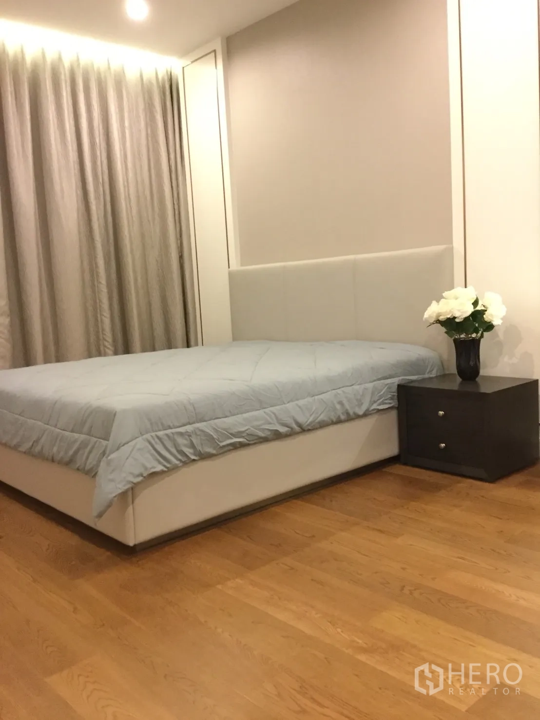 Condominium for rent in Sathon, Bangkok - Cozy bedroom with upholstered headboard, bedside table, full curtains and warm wood flooring.