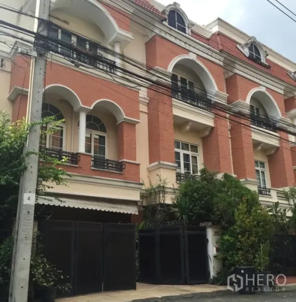 Townhouse for rent or sale in Bueng Kum, Bangkok - Three-storey townhouse exterior with red-brick facade, arched windows, balconies and a gated entrance framed by greenery.