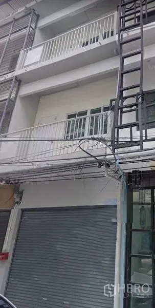 Shophouse for rent in Khlong Toei, Bangkok - Four-storey shophouse facade with roller shutter shopfront, metal-railed balconies, grey-white exterior and visible street wiring.