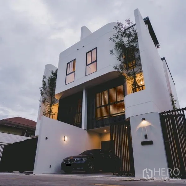 Townhouse for sale in Phra Khanong, Bangkok - Three-storey modern white townhouse with black slatted gate and a covered carport at dusk.
