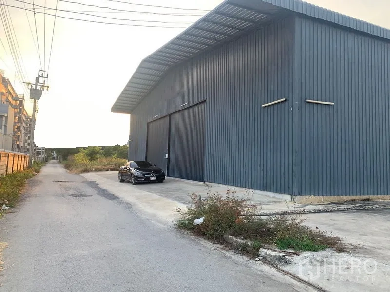 Warehouse for rent in Khan Na Yao, Bangkok - Side exterior of long metal warehouse along a quiet street with parking apron and power lines.