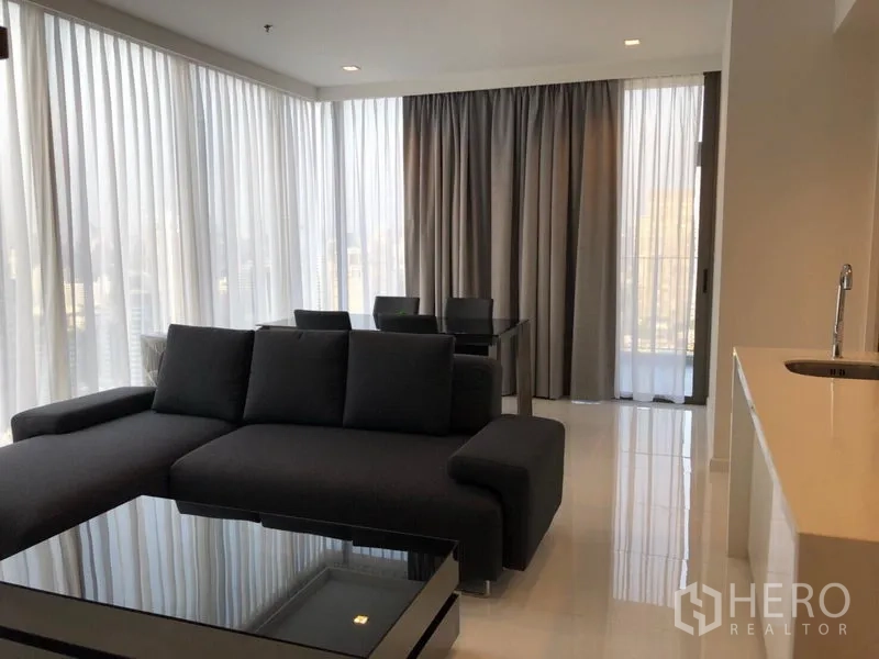 Condominium for rent in Sathon, Bangkok - Open‑plan corner lounge with L‑shaped sofa, dining set by large windows and a sliding door to the balcony.