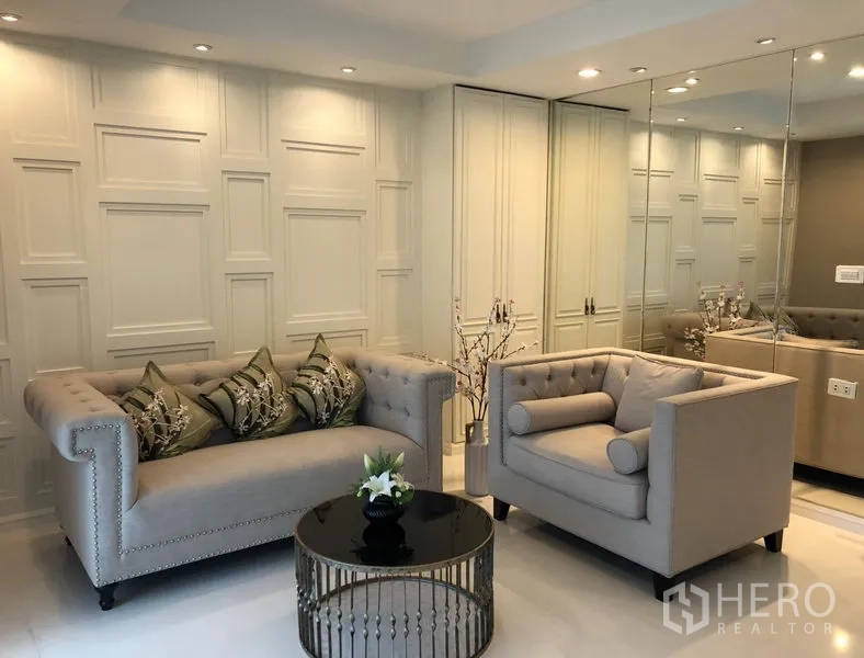 Condominium for rent or sale in Sathon, Bangkok - Elegant living room with tufted sofa and armchair, mirrored wall, round coffee table and recessed lighting in a Sathon Bangkok condo.