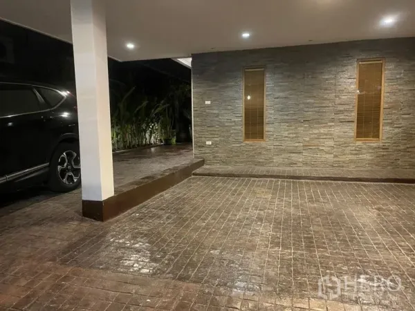 Townhouse for rent in Suan Luang, Bangkok - Covered carport with stone-clad wall, tiled driveway and recessed lighting beside greenery.