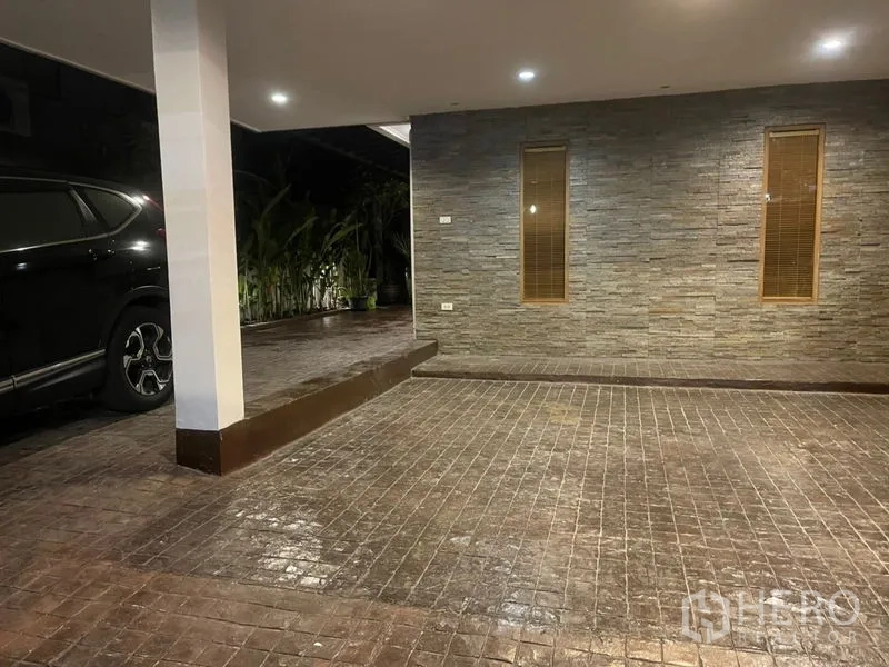 Townhouse for rent in Suan Luang, Bangkok - Covered carport with stone-clad wall, tiled driveway and recessed lighting beside greenery.