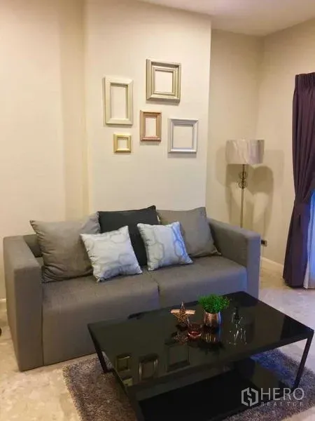 Condominium for rent in Khlong Toei, Bangkok - Cozy living room with a gray sofa, cushions, glass coffee table, neutral walls and a floor lamp.