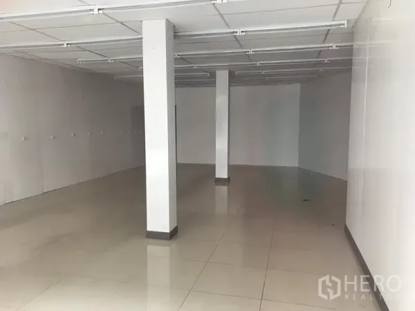 Retail Space for rent in Khlong Toei, Bangkok - Empty ground-floor retail unit with white walls, glossy tiled floor, support columns and a suspended grid ceiling with fluorescent light tracks.