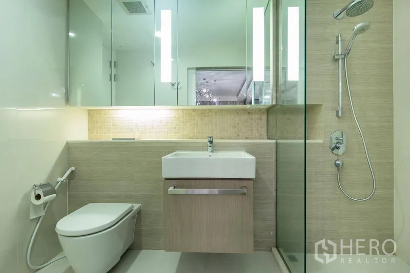 Condominium for rent in Watthana, Bangkok - Sleek bathroom with glass-enclosed shower, wall-hung toilet, vanity sink, and backlit mirror.
