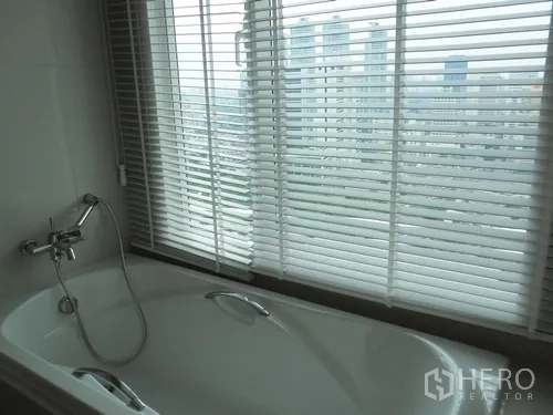 Condominium for rent or sale in Watthana, Bangkok - Bathroom with window-side bathtub and blinds overlooking the city.