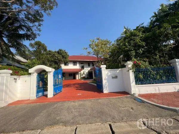 独立式住宅 出租在Bang Phli, Samut Prakan - 蓝色铁艺大门与白色围墙，通往带红色铺装车道与绿树的两层独栋住宅。