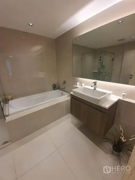 Condominium for rent in Khlong Toei, Bangkok - Modern bathroom with a soaking bathtub, floating vanity and large mirror in neutral tiles.
