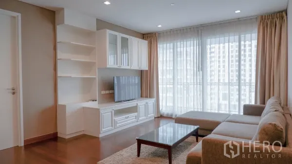 公寓 出租在Watthana, Bangkok - 明亮客厅，L形沙发、白色电视柜与落地窗及薄纱窗帘。