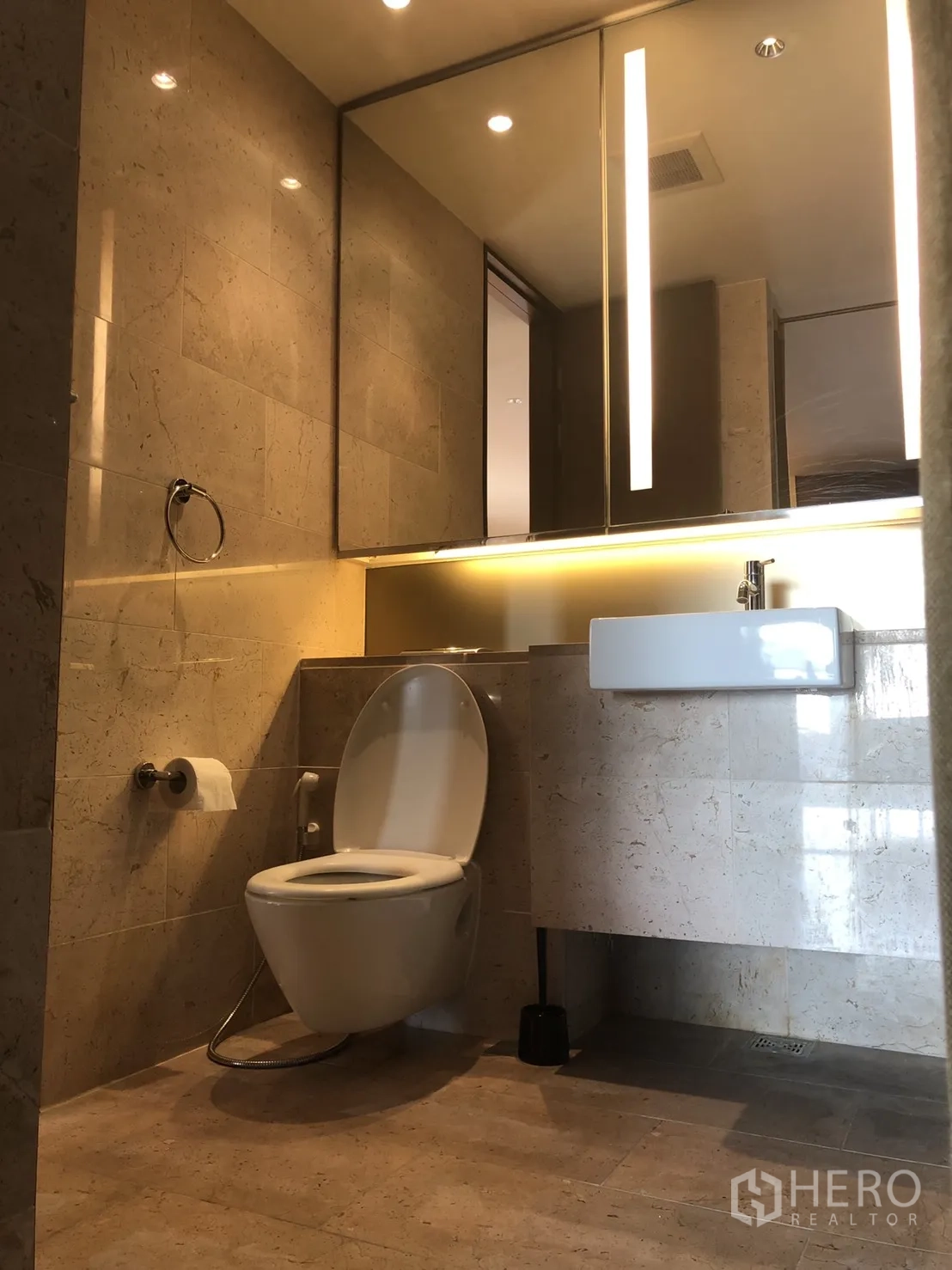 Condominium for rent in Pathum Wan, Bangkok - Modern bathroom with stone tiles, illuminated mirror, vanity sink, and contemporary toilet.