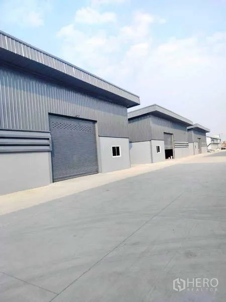 货栈 出租在Pathum Thani - 现代灰色金属仓库成排分布，配卷闸门和宽阔混凝土车道。